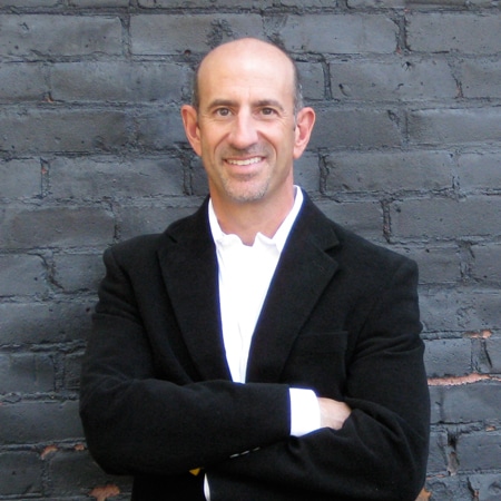 A man in a black blazer and white shirt stands with arms crossed in front of a dark brick wall. He is smiling and has a bald head.
