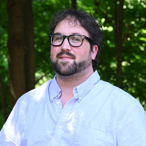 A man with glasses and a beard, wearing a light blue button-up shirt, stands in a wooded area. He looks slightly up and to the side. The background is filled with green leaves and tree trunks, suggesting a sunny day.