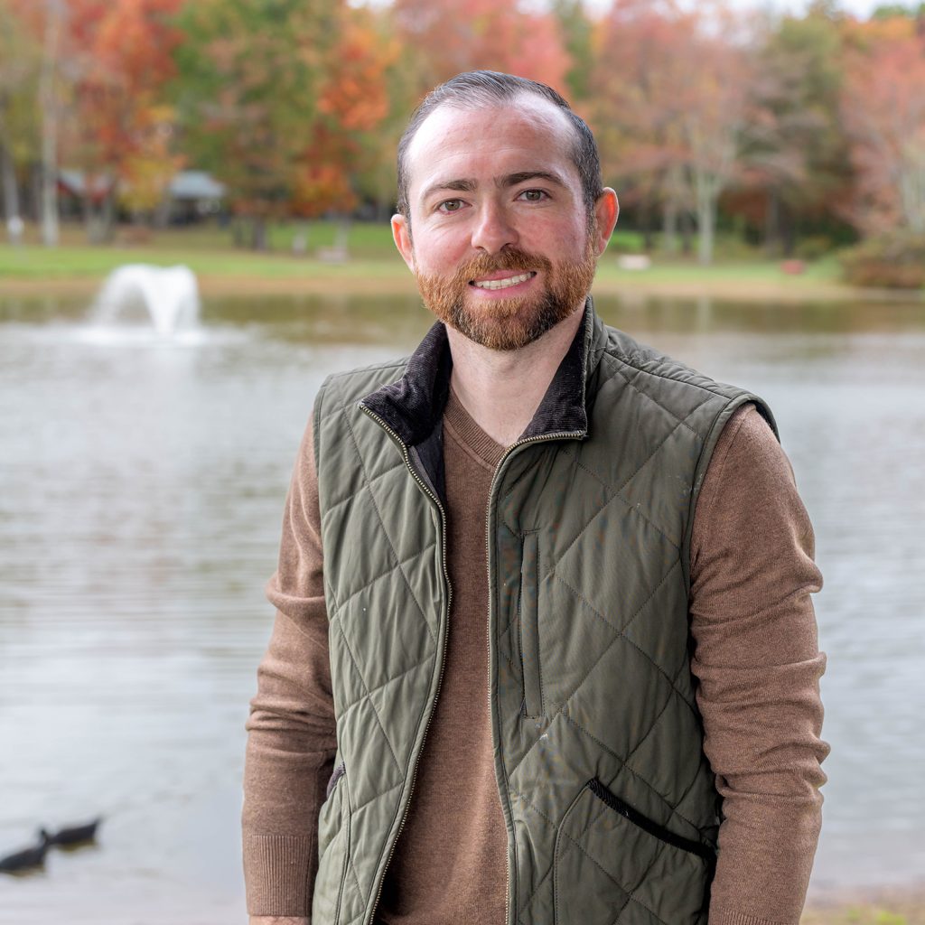 A man with short brown hair and a beard, wearing a green quilted vest over a brown sweater, stands by a lake with a fountain and autumn trees in the background.