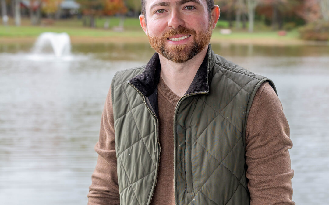 A man with short brown hair and a beard, wearing a green quilted vest over a brown sweater, stands by a lake with a fountain and autumn trees in the background.