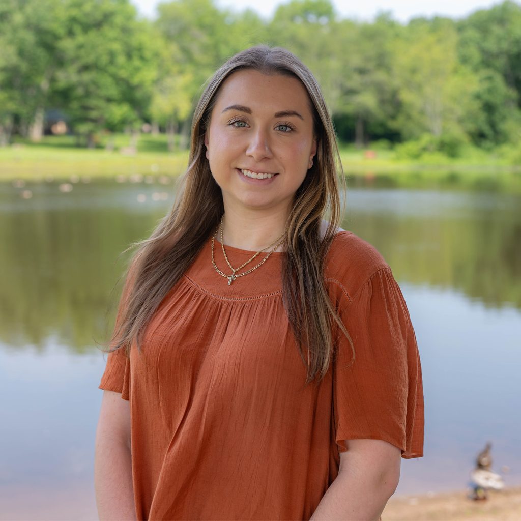 A young woman with long brown hair wearing an orange blouse stands smiling in front of a calm lake, with green trees and a grassy area visible in the background.