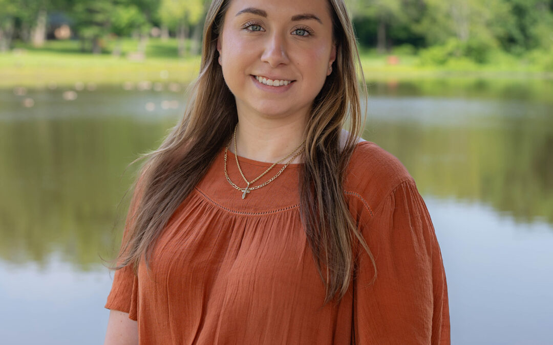 A young woman with long brown hair wearing an orange blouse stands smiling in front of a calm lake, with green trees and a grassy area visible in the background.
