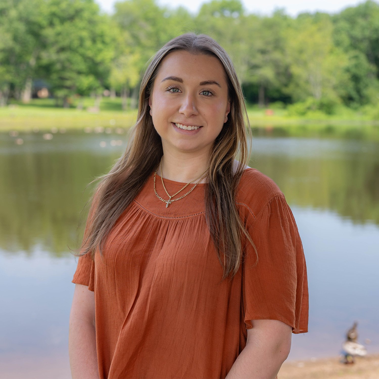A young woman with long brown hair wearing an orange blouse stands smiling in front of a calm lake, with green trees and a grassy area visible in the background.