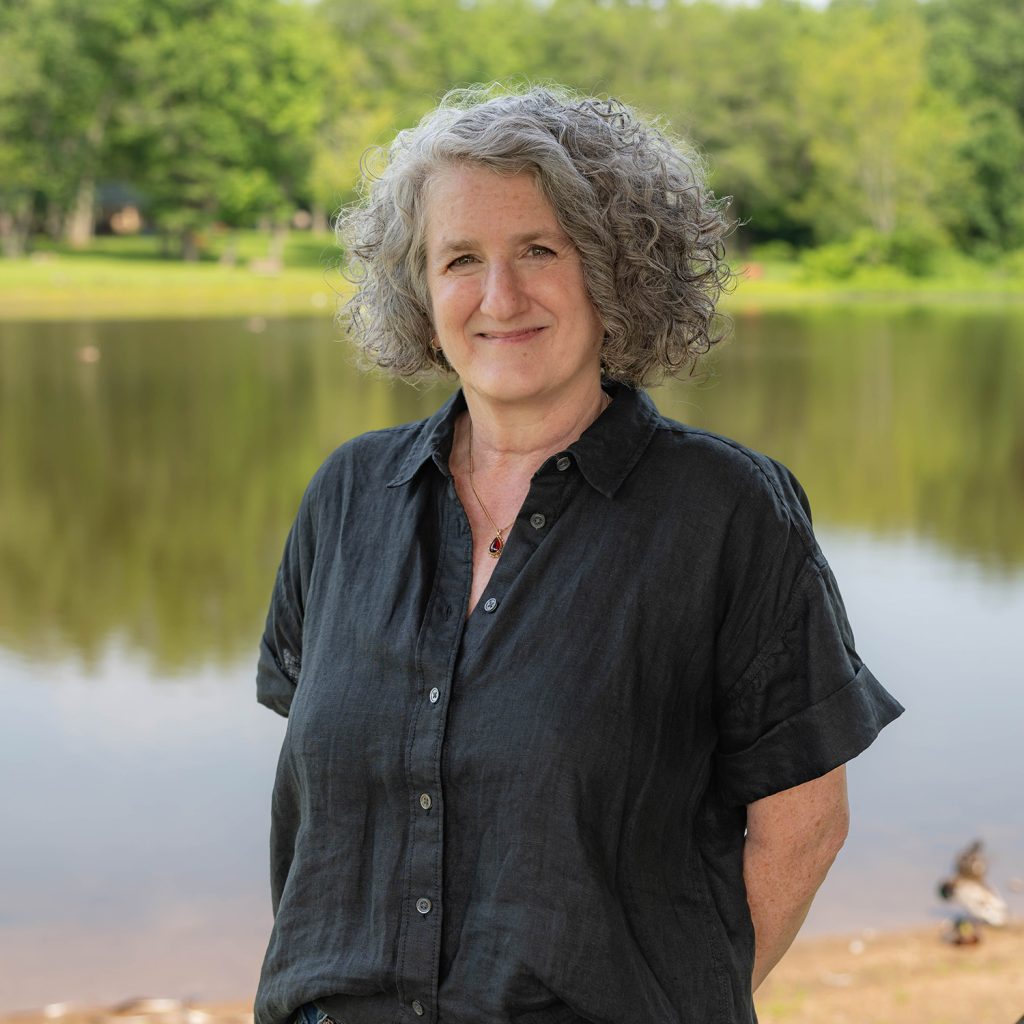 A woman with curly gray hair and a black button-up shirt stands smiling in front of a calm lake, with green trees and a grassy shore in the background.
