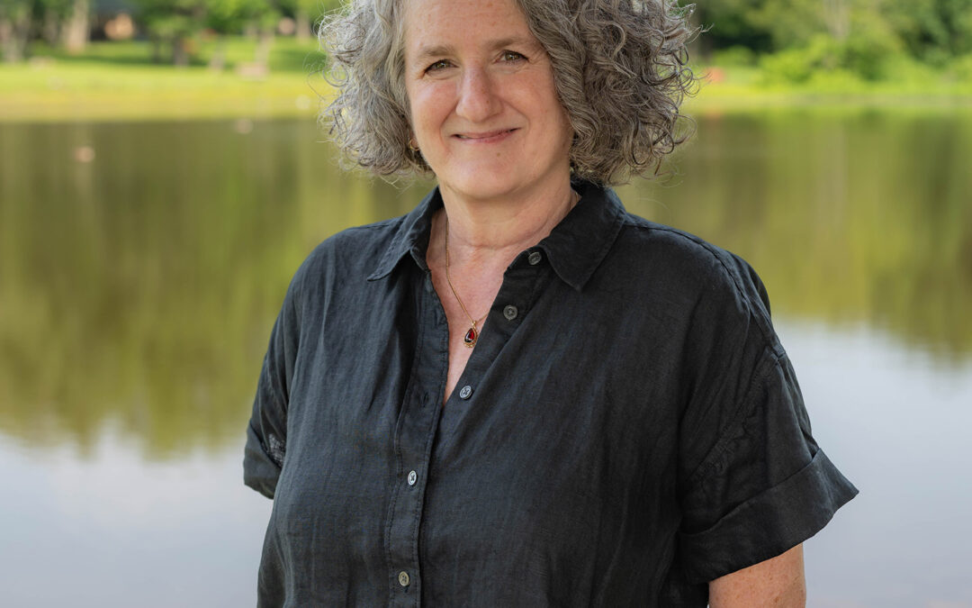 A woman with curly gray hair and a black button-up shirt stands smiling in front of a calm lake, with green trees and a grassy shore in the background.