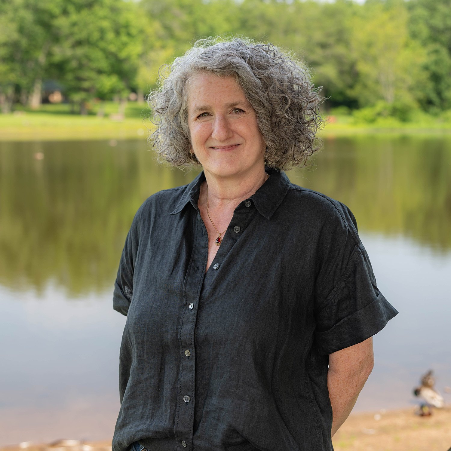 A woman with curly gray hair and a black button-up shirt stands smiling in front of a calm lake, with green trees and a grassy shore in the background.