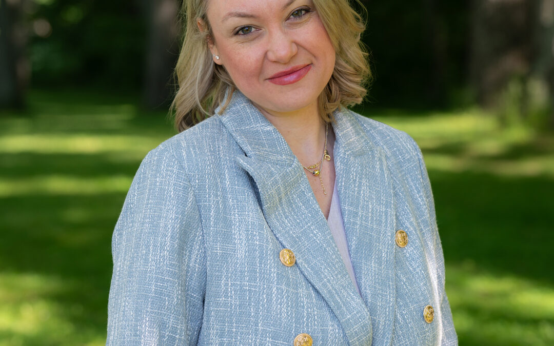 A woman with blonde hair, wearing a light blue blazer with gold buttons, stands outdoors on green grass with trees in the background, smiling gently at the camera.