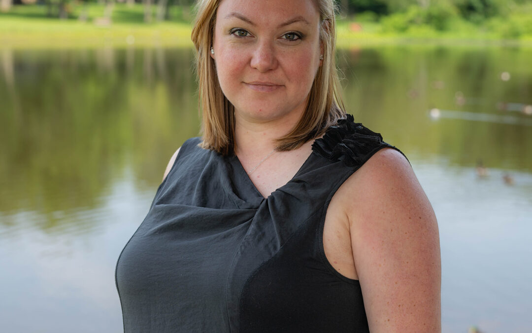 A woman with blonde hair wearing a sleeveless black top stands in front of a calm lake with trees and greenery in the background. She looks directly at the camera with a neutral expression.