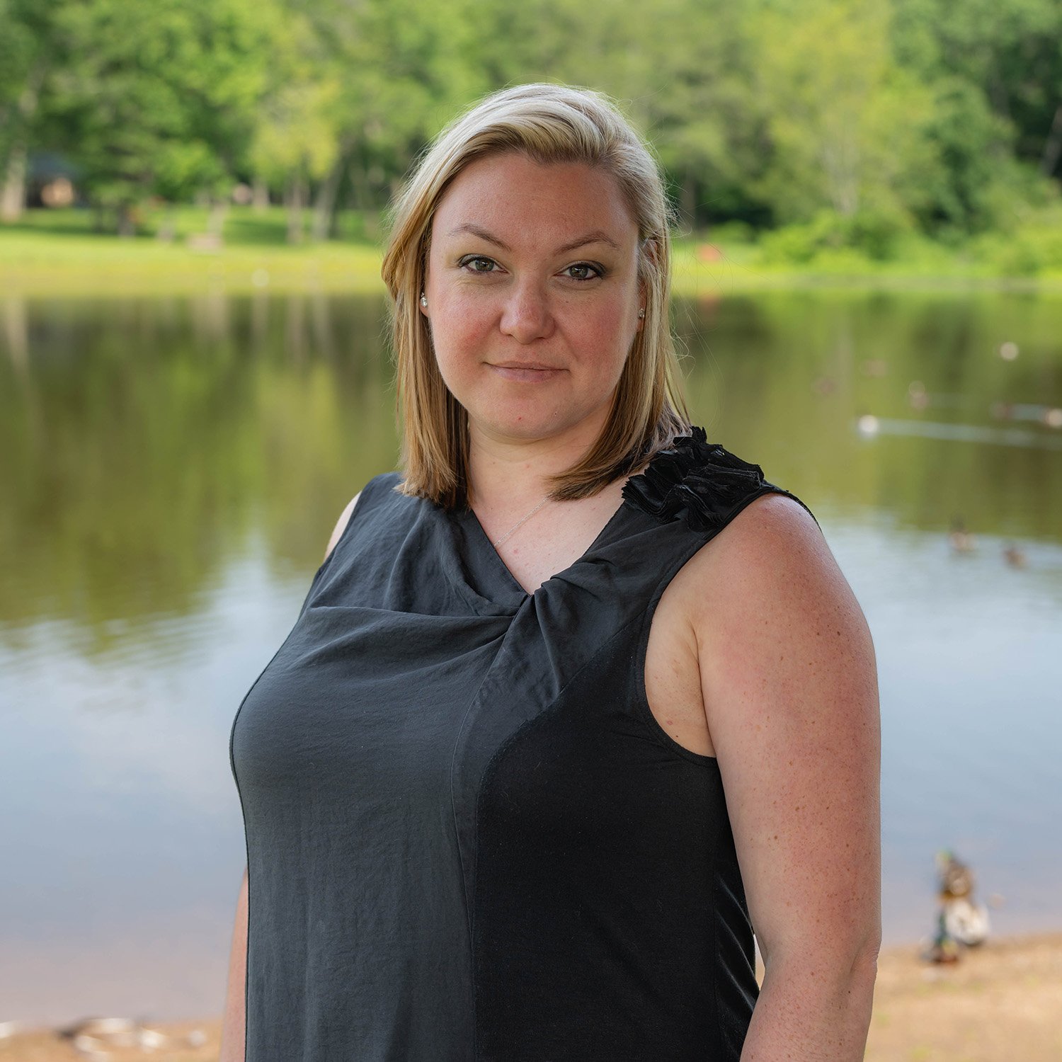 A woman with blonde hair wearing a sleeveless black top stands in front of a calm lake with trees and greenery in the background. She looks directly at the camera with a neutral expression.