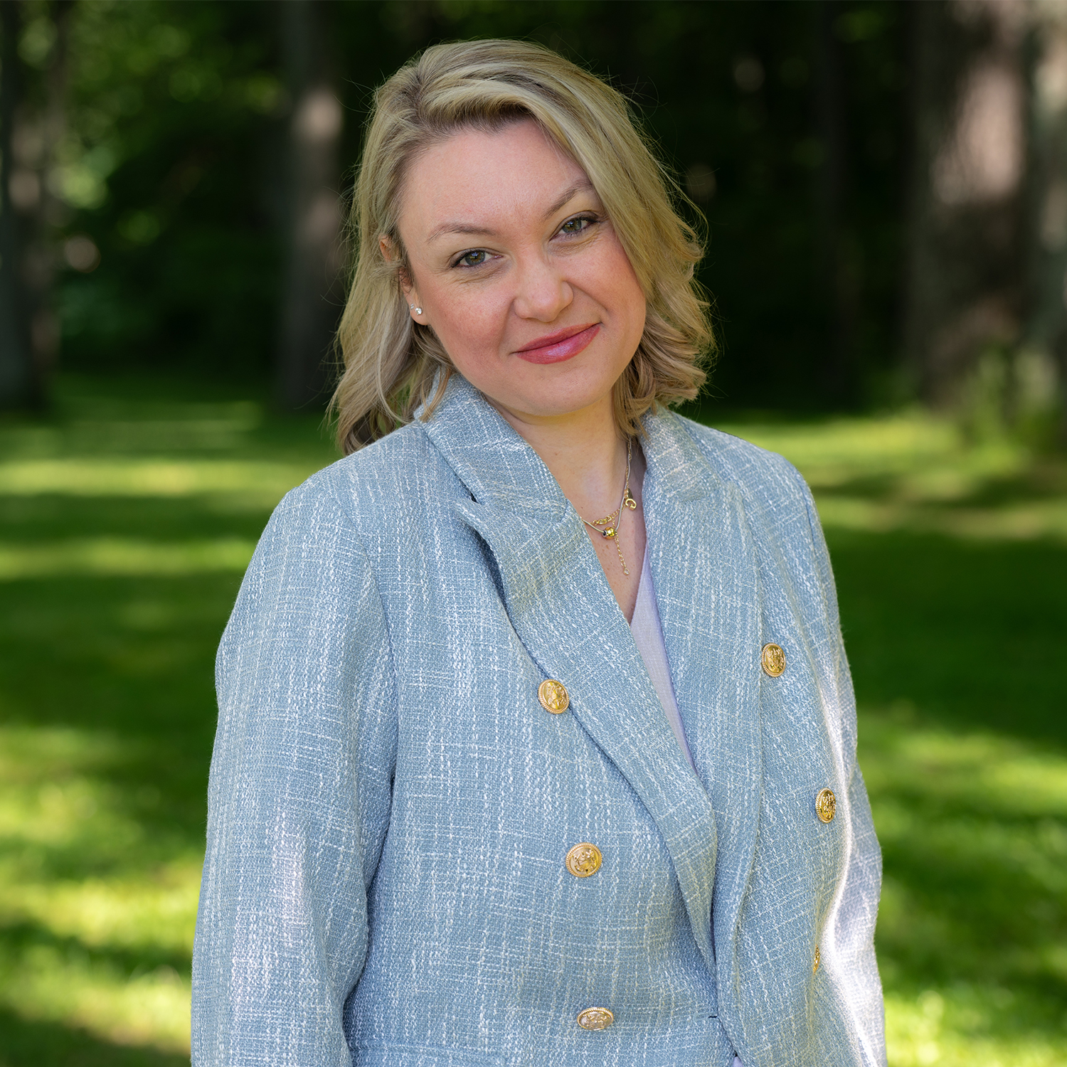 A woman with blonde hair, wearing a light blue blazer with gold buttons, stands outdoors on green grass with trees in the background, smiling gently at the camera.