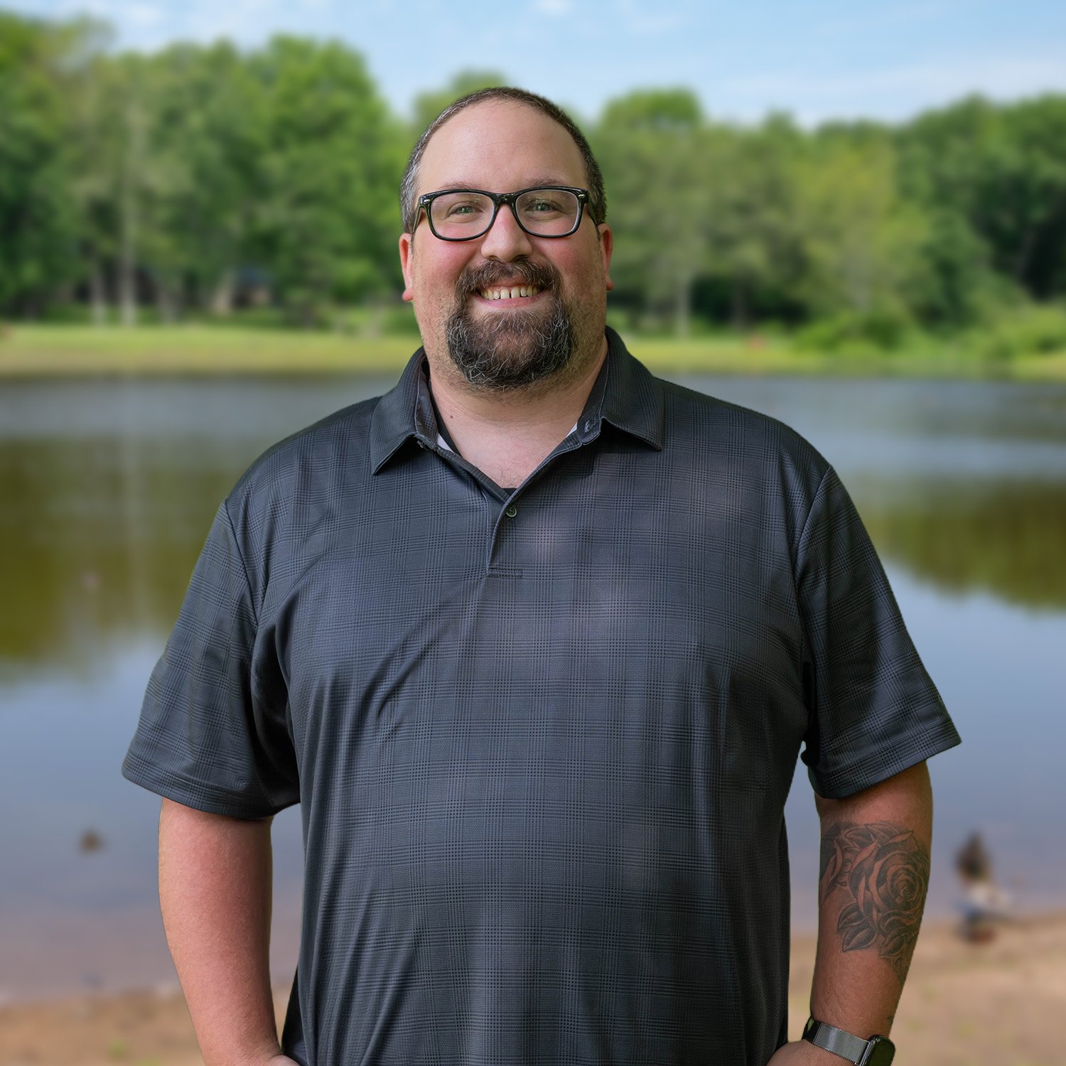 A smiling man with glasses and a beard stands outdoors in front of a calm lake, surrounded by green trees. He wears a dark short-sleeved polo shirt and has a tattoo on his right forearm.