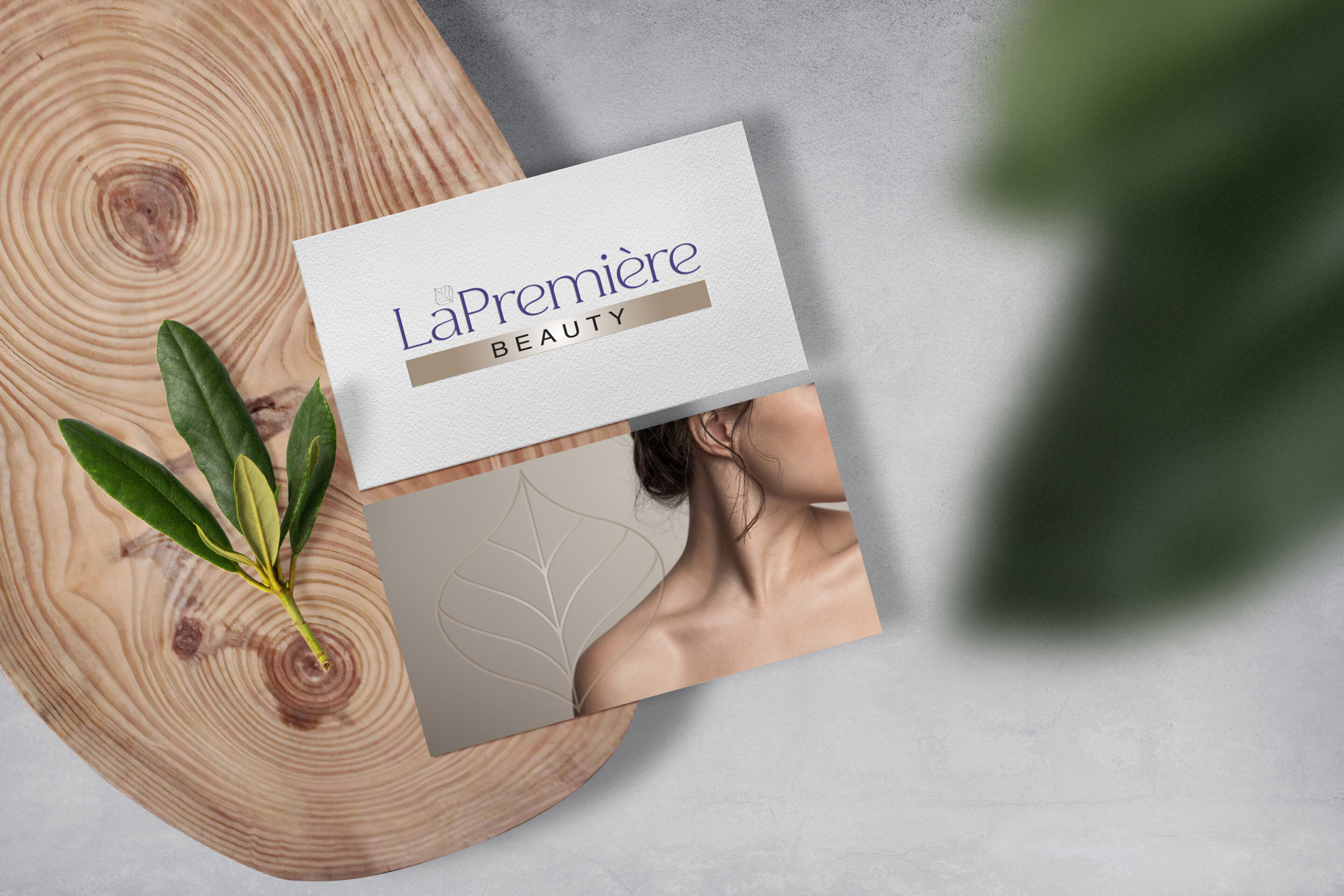 Two business cards on a wooden tray: one shows the “La Première Beauty” logo and the other has a partial view of a womans face and shoulder with a leaf outline design. A green leafy twig lies beside the cards.