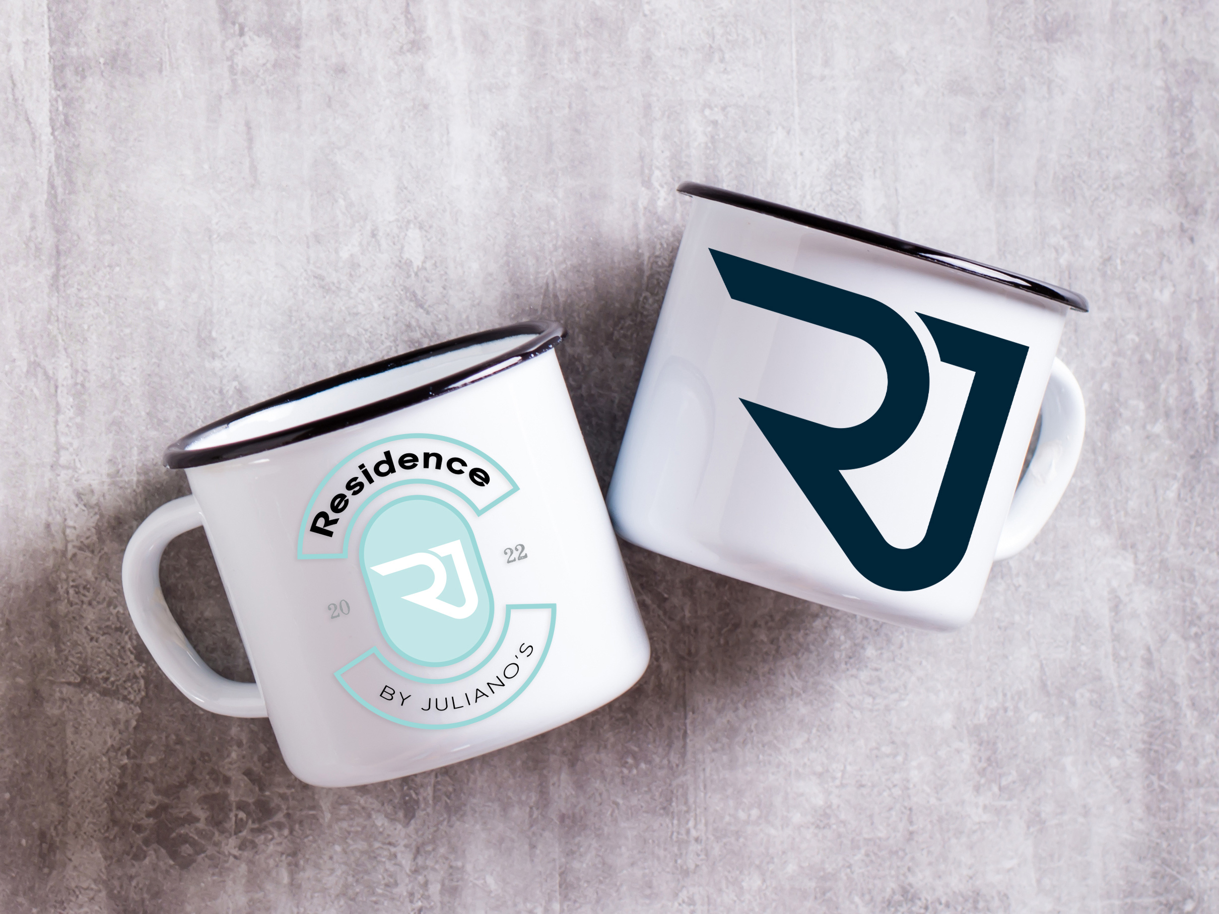 Residence-mugs Two white metal mugs on a gray surface, one lying down and one upright. Both feature a bold RJ logo; the left mug also has text reading Residence by Juliano’s with 2022 on either side of the logo.