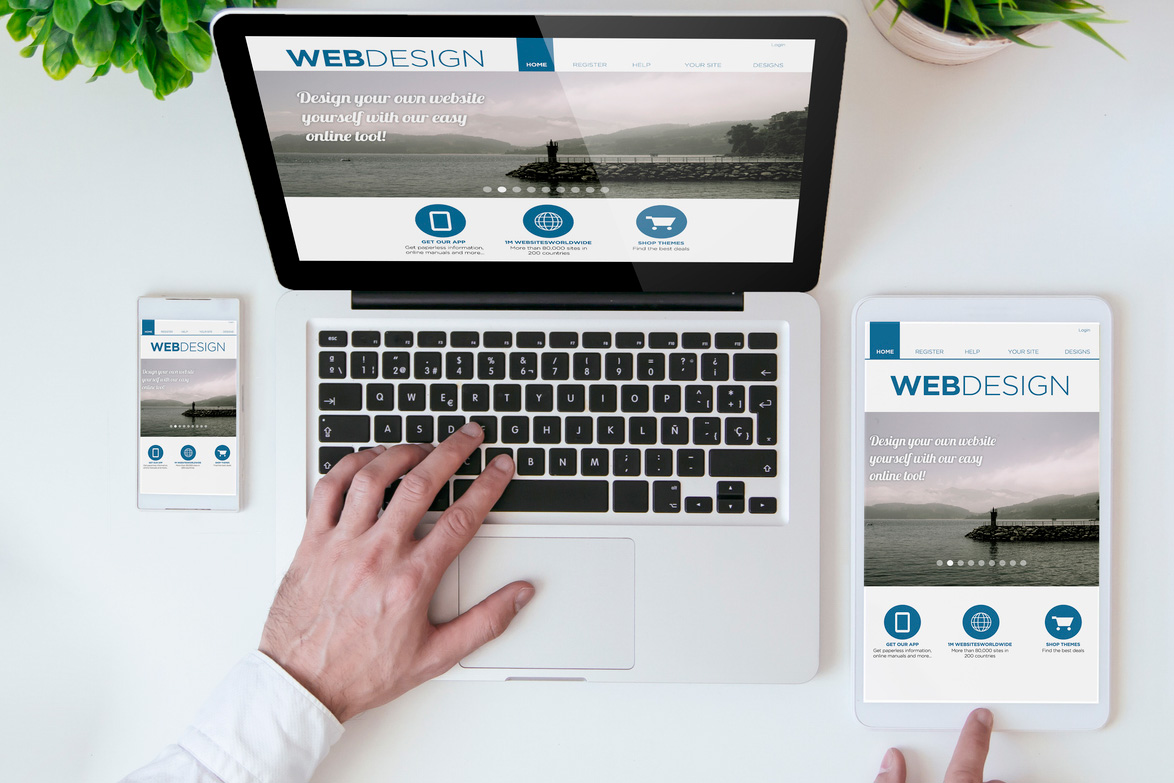 A person uses a laptop, with a smartphone and a tablet next to it, all displaying the same web design interface. The devices are on a white desk with a potted plant in the corner.