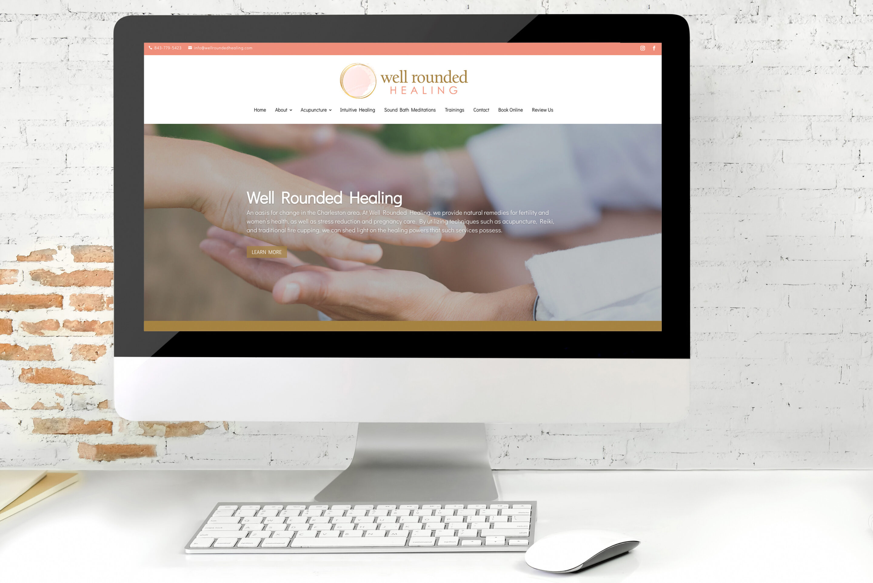 A desktop computer displays the Well Rounded Healing website, featuring an image of two hands reaching toward each other and text about holistic healing. The computer sits on a white desk against a brick wall.