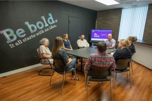 Eight people sit in a meeting room around a rectangular table, engaged in discussion. The wall behind them displays the phrase Be bold. It all starts with an idea. A screen shows a similar message.