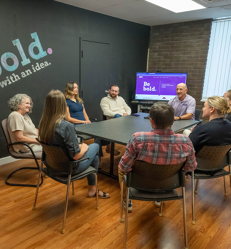 Eight people sit around a conference table in an office, engaged in a meeting. A screen displays a purple slide that reads “Be bold.” The wall behind them also features the words “bold.” and “with an idea.”.