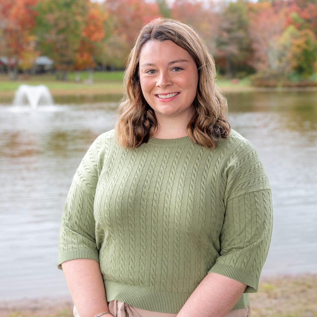 A woman with wavy brown hair wearing a light green sweater stands smiling in front of a lake with a water fountain and colorful autumn trees in the background.