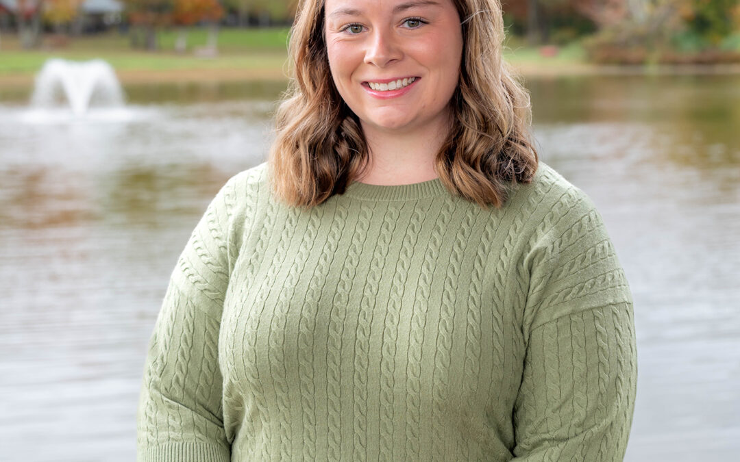 A woman with wavy brown hair wearing a light green sweater stands smiling in front of a lake with a water fountain and colorful autumn trees in the background.
