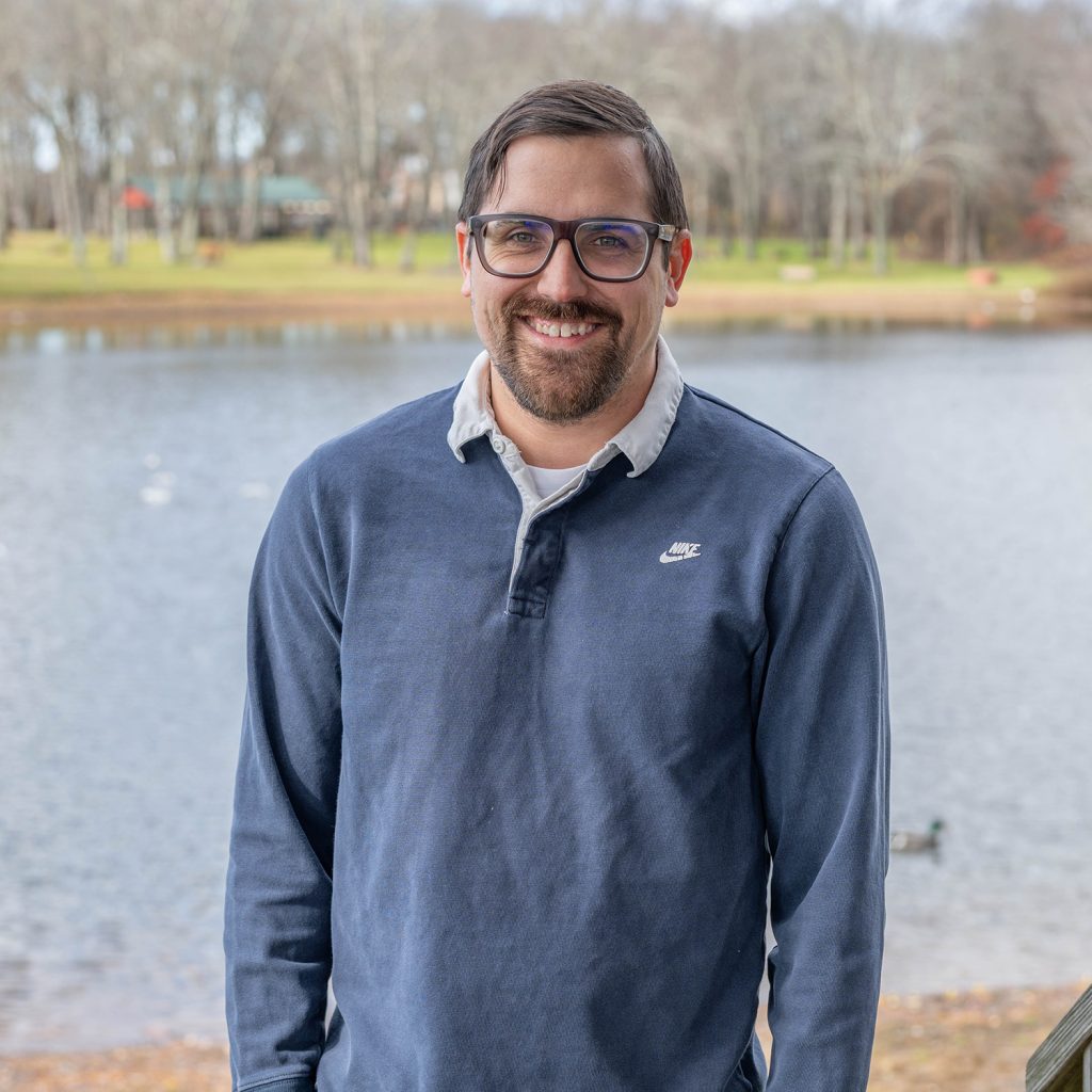 A man with glasses, short dark hair, and a beard is smiling while standing in front of a lake with trees and grass in the background. He wears a blue Nike pullover over a collared shirt.