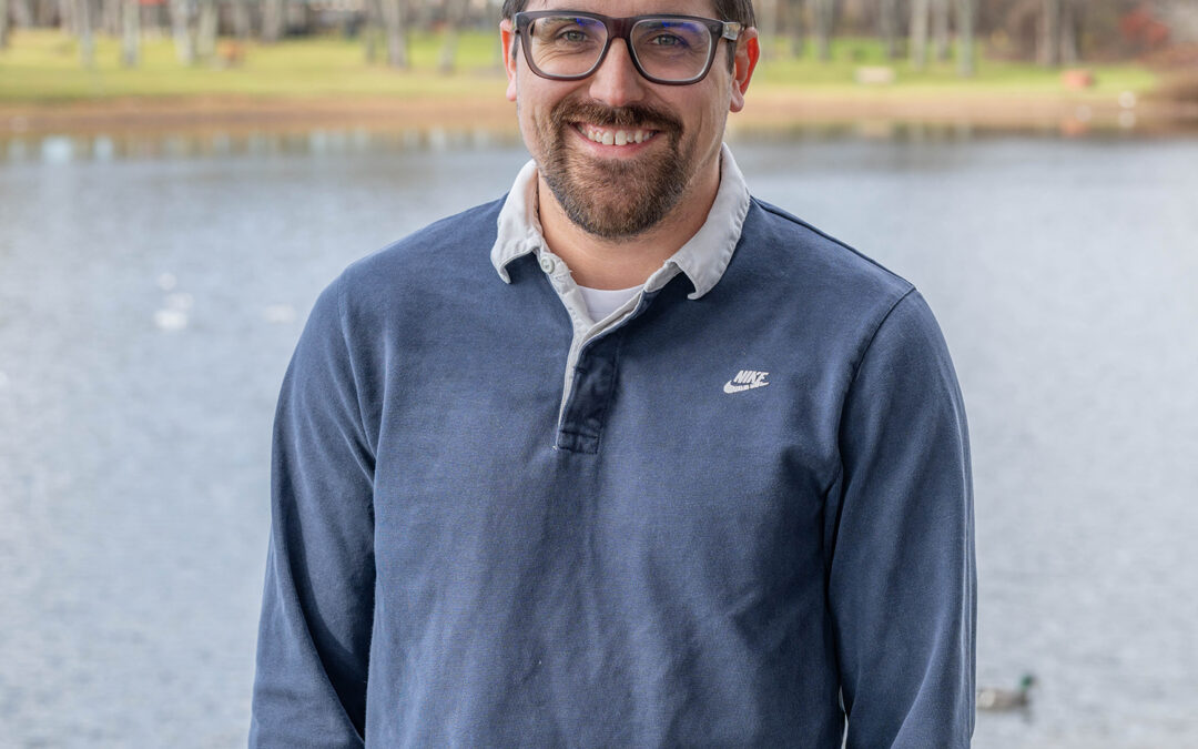 A man with glasses, short dark hair, and a beard is smiling while standing in front of a lake with trees and grass in the background. He wears a blue Nike pullover over a collared shirt.