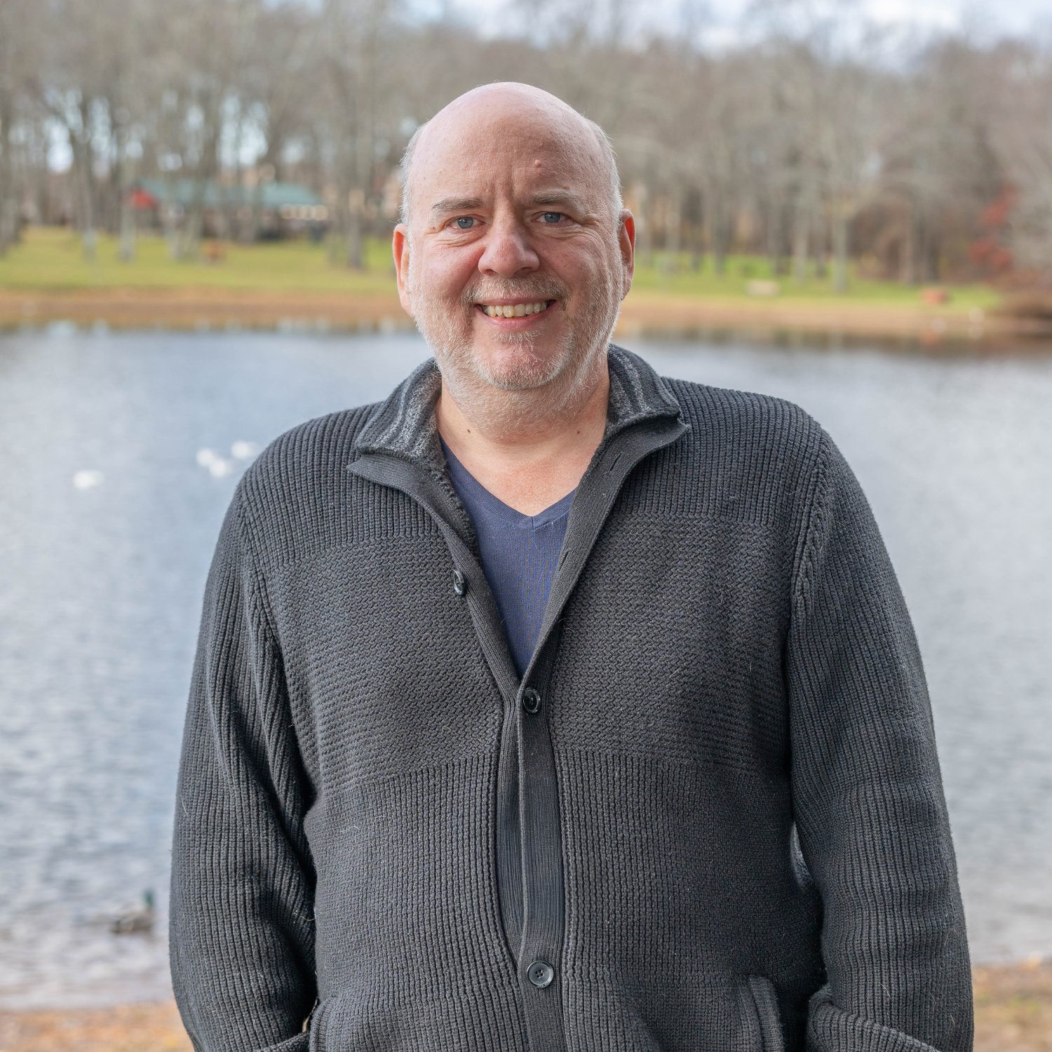 A middle-aged man with a light beard is smiling while standing outdoors in front of a lake, wearing a dark gray sweater over a navy shirt. Bare trees and a grassy area are visible in the background.