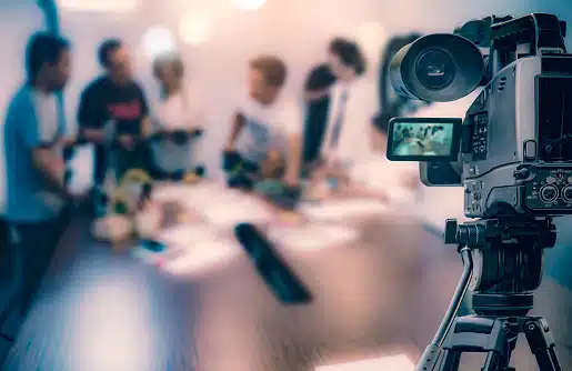 A video camera on a tripod is focused on a group of people having a discussion around a conference table, with the scene blurred in the background.