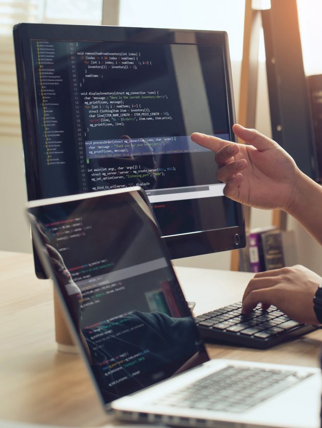 Developing programming and coding technologies. Professional Developer programmer cooperation meeting brainstorming and analysis programming in website working and coding technology.Website design. Two people sit at a desk, looking at a computer monitor with code displayed. One person points at the screen while the other watches. A laptop with code on its screen is also on the desk next to them.