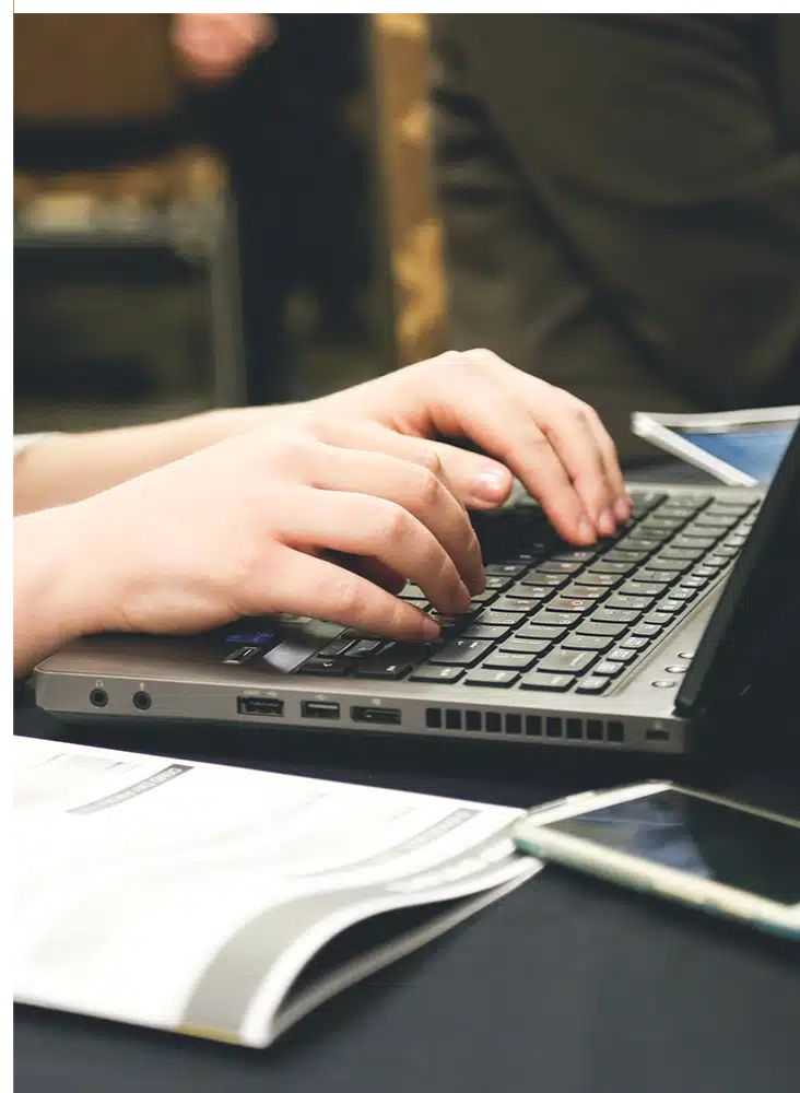 envision-marketing-laptop-inuse-ct A person types on a laptop keyboard with both hands. An open notebook or document and a smartphone are placed nearby on the table.