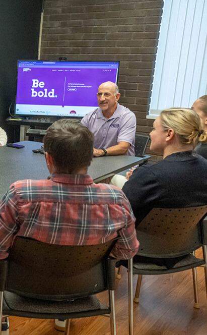 envision-marketing-team-web-design-ct-2 Four people sit around a conference table having a discussion, with a man at the head of the table. A monitor in the background displays a purple screen with the words Be bold.