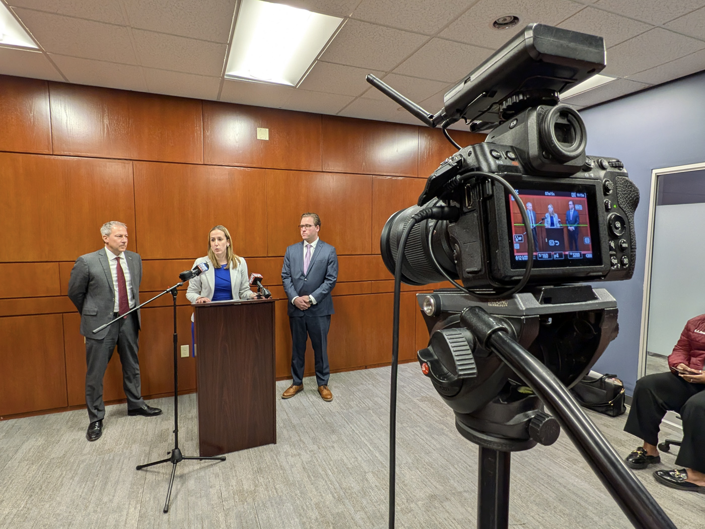 A woman stands at a podium speaking into microphones, flanked by two men in suits. A video camera in the foreground records the scene. The setting appears to be a conference room with wood-paneled walls.