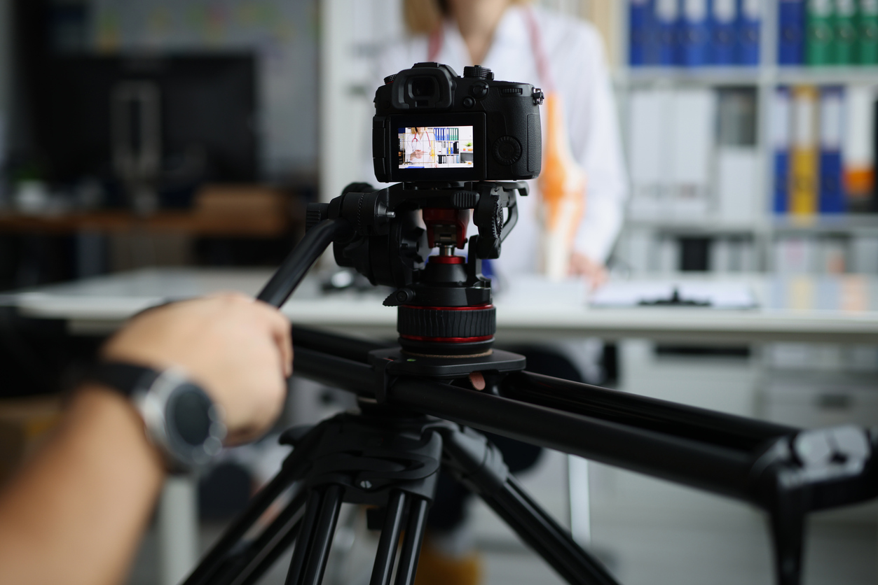 A person operates a camera on a tripod, filming someone sitting at a desk in an office with shelves and binders in the background. The filmed person is out of focus.