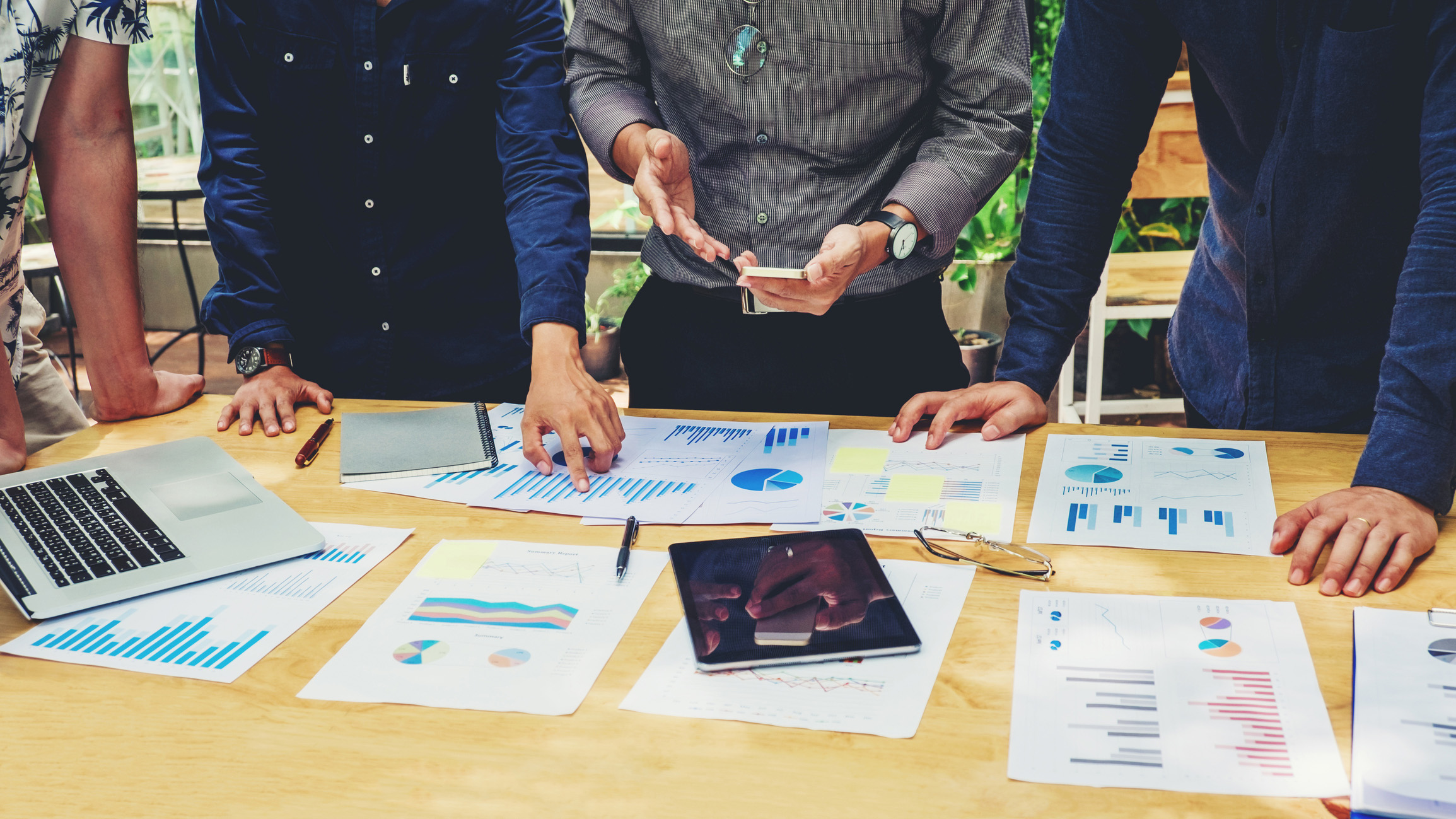 SEO vs Social Media for my business Four people stand around a table covered with charts, graphs, documents, a laptop, and a tablet. One person gestures with their hands while others look at the papers, suggesting a business meeting or discussion.