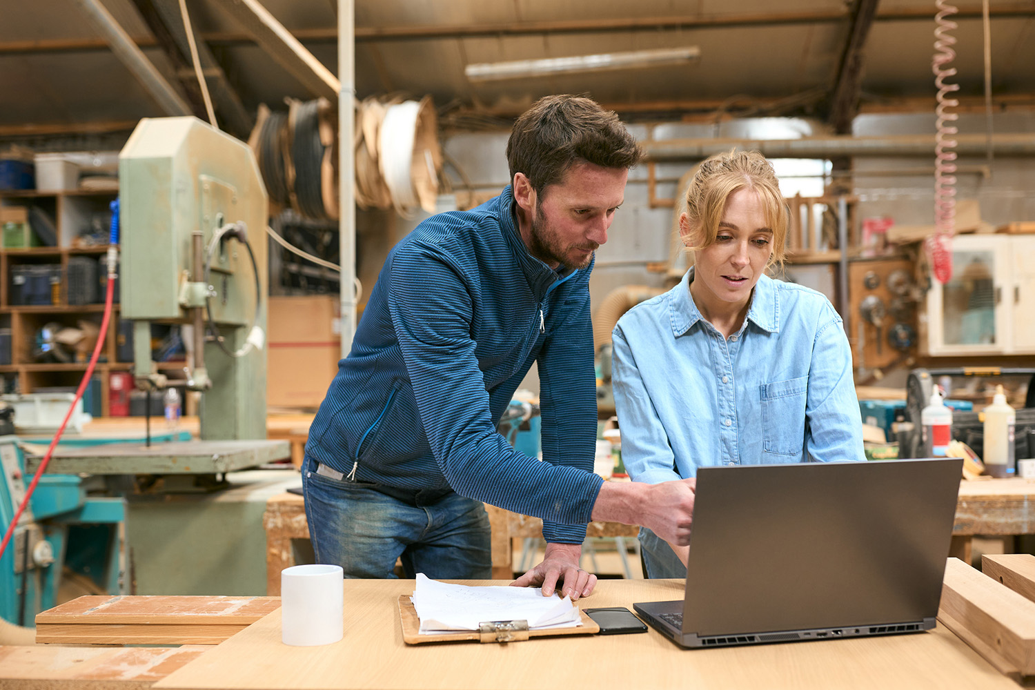 A man and a woman in a woodworking shop look at a laptop on a workbench. The man is pointing at the screen, and both appear focused. Tools, wood, and equipment are visible in the background.