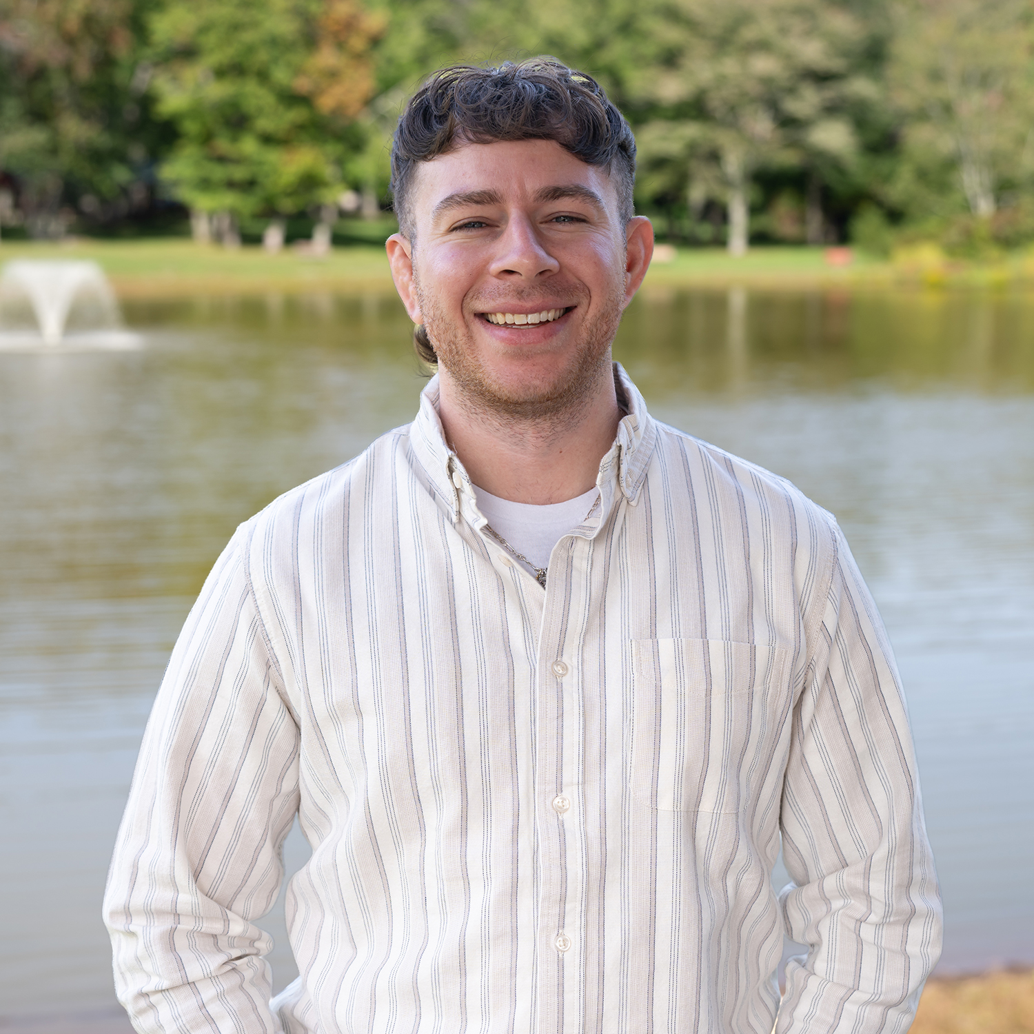 A smiling person in a striped shirt stands in front of a pond with a fountain and trees in the background on a sunny day.