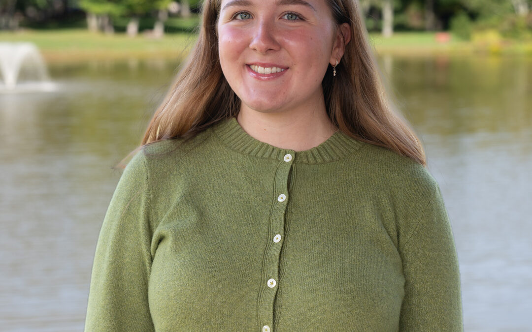 A woman with long light brown hair, wearing a green buttoned sweater, stands and smiles in front of a pond with a water fountain and trees in the background.