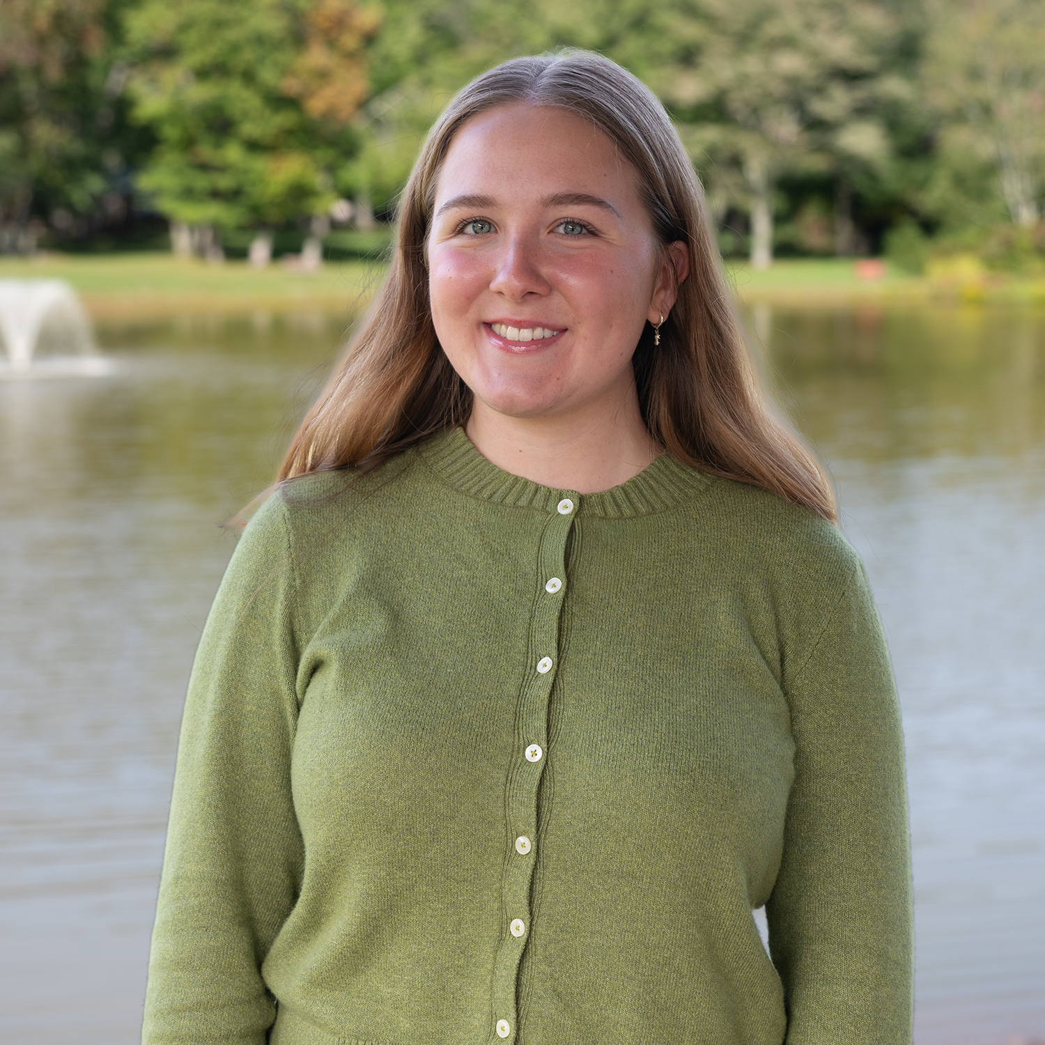 A woman with long light brown hair, wearing a green buttoned sweater, stands and smiles in front of a pond with a water fountain and trees in the background.